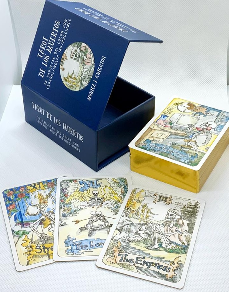 Pode incluir: Uma caixa azul com detalhes dourados contendo um baralho de cartas de tar&ocirc; com ilustra&ccedil;&otilde;es de esqueletos. A caixa tem o texto "Tarot de los Muertos" e "78 Tarjetas del Color con Espa&ntilde;ol/Ingles Instrucciones" na frente. As cartas est&atilde;o dispostas sobre uma superf&iacute;cie branca, mostrando as cartas "For&ccedil;a", "Os Amantes" e "A Imperatriz".