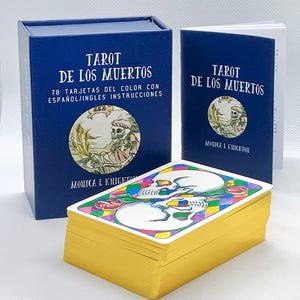 Pode incluir: Uma caixa azul com o texto "Tarot de los Muertos" e "78 Tarjetas del Color con Espa&ntilde;ol/Ingles Instrucciones". A caixa cont&eacute;m um baralho de cartas de tar&ocirc; com ilustra&ccedil;&otilde;es coloridas de caveiras e flores. As cartas est&atilde;o empilhadas em cima de uma caixa azul menor com o texto "Tarot de los Muertos" e "Monica L Knighton".