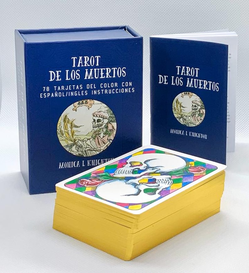 Pode incluir: Uma caixa azul com o texto "Tarot de los Muertos" e "78 Tarjetas del Color con Espa&ntilde;ol/Ingles Instrucciones". A caixa cont&eacute;m um baralho de cartas de tar&ocirc; com ilustra&ccedil;&otilde;es coloridas de caveiras e flores. As cartas est&atilde;o empilhadas em cima de uma caixa azul menor com o texto "Tarot de los Muertos" e "Monica L Knighton".