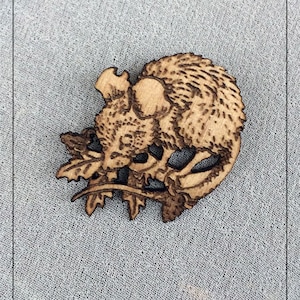 May include: A wooden squirrel-shaped brooch with intricate etched details. The brooch features a squirrel perched on a branch with leaves, showcasing a natural wood grain and a burnt-wood effect for added dimension. The brooch is set against a light gray background.