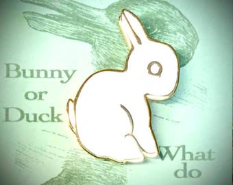 Pode incluir: Um alfinete de ilusão de óptica de coelho ou pato branco com acabamento dourado. O alfinete está sobre um fundo verde com o texto "Bunny or Duck" e "What do you see?"