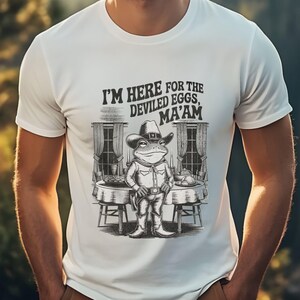 May include: White t-shirt with a black and white illustration of a frog wearing a cowboy hat and holding a gun. The frog is sitting at a table with food on it. The text on the shirt reads "I'm here for the deviled eggs, ma'am."