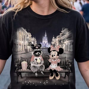 Może przedstawiać: Czarna koszulka z grafiką szopa pracza i Myszki Minnie siedzących na ławce, jedzących popcorn, z zamkiem i sceną uliczną w tle. Szop nosi okulary przeciwsłoneczne i kapelusz.