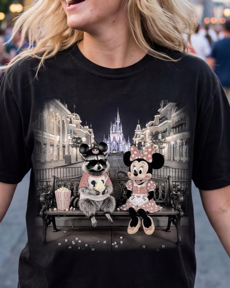 Może przedstawiać: Czarna koszulka z grafiką szopa pracza i Myszki Minnie siedzących na ławce, jedzących popcorn, z zamkiem i sceną uliczną w tle. Szop nosi okulary przeciwsłoneczne i kapelusz.