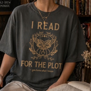 May include: Dark gray t-shirt with the text "I READ FOR THE PLOT" in gold lettering. A gold illustration of a moth, moons, and crystals is centered below the text. The phrase "- you know what I mean" is printed below the main text.