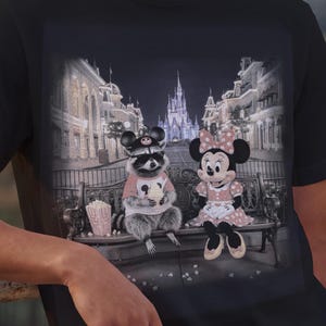 Może przedstawiać: Czarny t-shirt z fantazyjną grafiką szopa pracza i Myszki Minnie siedzących na ławce przed zamkiem. Szop nosi okulary przeciwsłoneczne i r&oacute;żową koszulkę, a Myszka Minnie ma na sobie r&oacute;żową sukienkę w kropki. W pobliżu znajduje się pudełko popcornu.