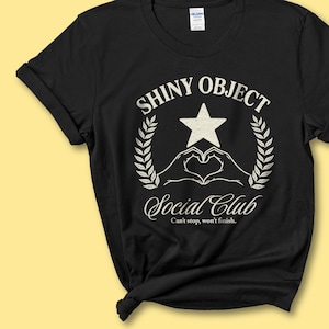 Puede incluir: Camiseta negra con un gráfico blanco que dice "SHINY OBJECT Social Club" con una estrella, manos formando un corazón y hojas de laurel. El texto "Can't stop, won't finish" está debajo del nombre del club.