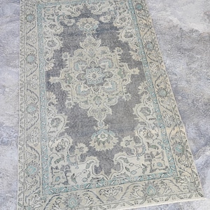 Puede incluir: Una alfombra vintage con fondo gris claro y un patrón floral beige y turquesa. La alfombra tiene un borde con flecos.