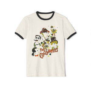 Puede incluir: Una camiseta blanca con cuello de canalé negro que presenta una ilustración vintage de un grupo de personas con el texto "The Cocoanuts" impreso en la parte delantera.