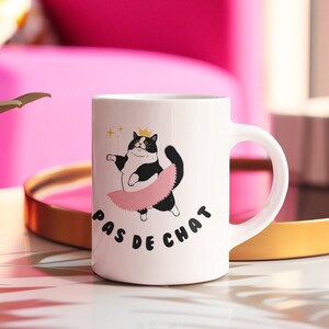 May include: White ceramic mug with a black and white cat wearing a pink tutu and a gold crown. The text "Pas de Chat" is printed on the mug.