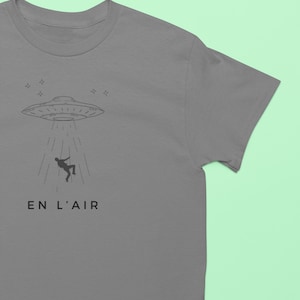 Puede incluir: Una camiseta gris con una ilustración en blanco y negro de un OVNI secuestrando a una persona. El texto "EN L'AIR" está impreso debajo de la ilustración.
