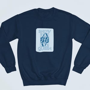 Op de afbeelding: Een donkerblauw sweatshirt met een witte afbeelding van een speelkaart. De kaart heeft de tekst "SNOW QUEEN" en toont een gestileerde afbeelding van een koningin die een zwaard vasthoudt.
