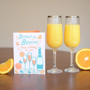 Może przedstawiać: Kartka urodzinowa z tekstem "Bitches be Brunchin' happy birthday to you" i ilustracją kieliszk&oacute;w do szampana i butelki szampana. W pierwszym planie znajdują się dwa kieliszki soku pomarańczowego.