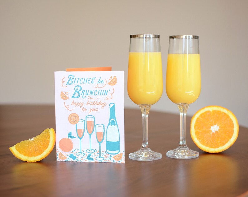 Może przedstawiać: Kartka urodzinowa z tekstem "Bitches be Brunchin' happy birthday to you" i ilustracją kieliszk&oacute;w do szampana i butelki szampana. W pierwszym planie znajdują się dwa kieliszki soku pomarańczowego.