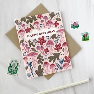 May include: A birthday card with a pink background and a pattern of mushrooms, flowers, and leaves. The words "HAPPY BIRTHDAY" are in a white banner. The card is on a brown envelope, with stamps and a pencil nearby.