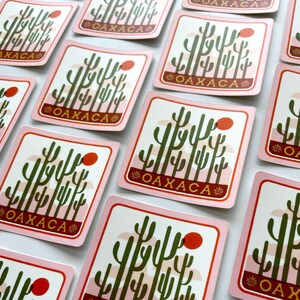 May include: A set of pink and white stickers featuring a graphic of a cactus landscape and the word "Oaxaca" in gold lettering. The stickers are square shaped and have rounded corners.