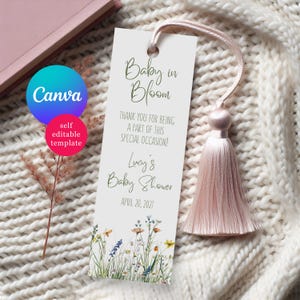 May include: A white bookmark with a pink tassel, featuring the text "Baby in Bloom" and "Lucy's Baby Shower, April 20, 2027." The bottom of the bookmark has a floral design with wildflowers. The Canva logo is visible.