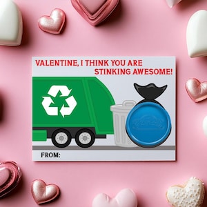 May include: Valentine's Day card with a green garbage truck illustration and the text "VALENTINE, I THINK YOU ARE STINKING AWESOME!". The card features a recycling symbol and a blue trash can lid. Surrounded by pink and white heart-shaped candies.