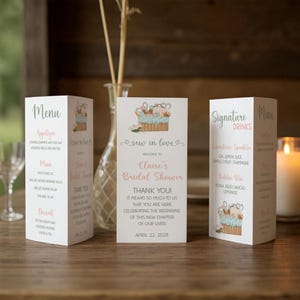 Puede incluir: Tres tarjetas de menú blancas y plegadas y una tarjeta central con texto para una despedida de soltera. Las tarjetas presentan un diseño floral y de costura. El texto incluye "Menú", "Bebidas de la firma" y "Despedida de soltera de Claire".