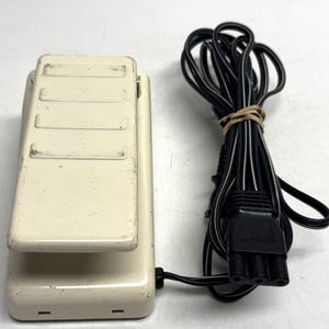 May include: Off-white sewing machine foot pedal with a black power cord. The pedal has a rectangular shape with three recessed areas on the top. The power cord is coiled and secured with a rubber band.