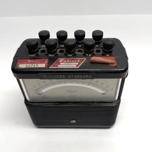 May include: A vintage black Babcock Electronics Corporation Transfer Standard voltmeter. The device has a series of black knobs on top, a gauge with a needle, and the Babcock logo. The text "VOLTS A.C." is visible on the gauge.