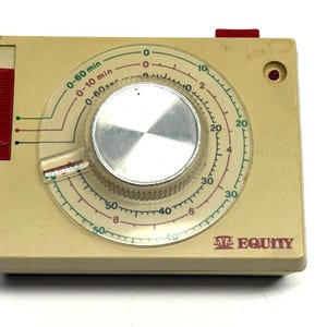 May include: A vintage cream-colored kitchen timer with a clear dial and a large silver knob. The dial displays time increments from 0 to 60 minutes, 0 to 10 minutes, and 0 to 60 seconds. It has a red button and a red side panel. The brand name "AVA EQUITY" is printed on the bottom.
