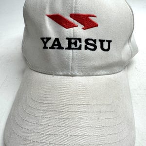 May include: White baseball cap with a curved brim. The cap features the word "YAESU" in black block letters, with a red logo above. The cap is made of a textured fabric.