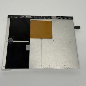 May include: A metal paper trimmer featuring a silver base with a black sliding guide, a black top ruler marked from 0 to 4 inches on both sides, and a square tan material piece positioned in the center of the silver surface for alignment.