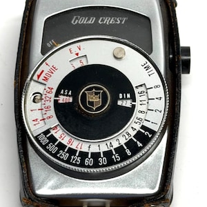 May include: A vintage Gold Crest light meter with a silver and black face, encased in a brown leather case. The meter features a dial with ASA, DIN, and time settings, along with the text "MOVIE".
