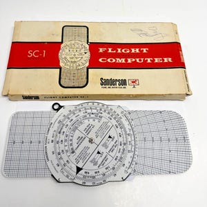 May include: Vintage Sanderson Flight Computer SC-1. The flight computer is white and black with a circular slide rule design. The box is beige with red accents and the words "Flight Computer" printed on it.