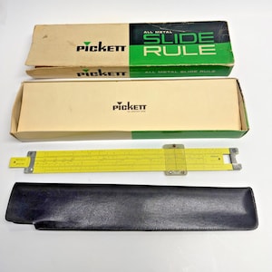 May include: A vintage Pickett slide rule with its original packaging and a black leather case. The slide rule is yellow with black markings and a clear cursor. The boxes are tan with green and black accents, and the Pickett logo.