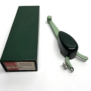 May include: An adjustable scriber tool with a dark green handle and a black grip, next to a dark green box. The box has a label that reads "Doric 613160 Adjustable Scriber". The tool is likely used for marking or measuring.