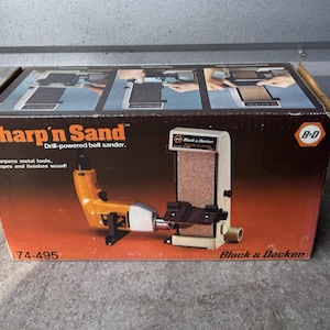 May include: A Black & Decker Sharp'n Sand drill-powered belt sander in its original box. The box is black with red and white text. The sander is a light beige color with a black belt and a black and yellow drill attachment. The box also includes the model number 74-495.