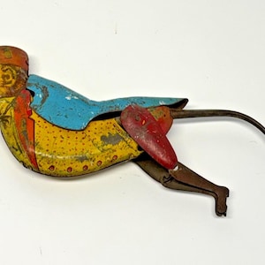 May include: A vintage tin wind-up toy shaped like a monkey wearing a red hat, a yellow jacket with red trim, and a blue cape. The figure has a long metal tail, articulated red limbs, and shows signs of paint wear and oxidation on the metal surface.
