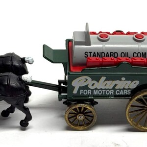 May include: A toy model of a horse-drawn oil tanker. The tanker is gray with red accents and the words "Standard Oil Company" and "Polarine for Motor Cars" printed on it. The horses are black with white manes and tails.
