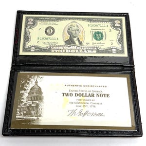 May include: A black leather wallet displaying a two-dollar bill. The bill features a portrait of Thomas Jefferson and the words "TWO DOLLARS." The wallet also contains a certificate that reads "AUTHENTIC UNCIRCULATED TWO DOLLAR NOTE."