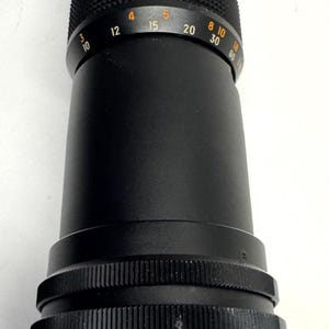 May include: A black camera lens with a ribbed focus ring and a series of numerical markings in orange and white. The lens has a cylindrical shape with a textured grip section and a lens mount at the base.