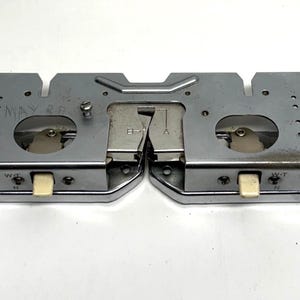 May include: A silver-colored metal sewing machine attachment with two rectangular sections and rounded edges. Each section has a small, off-white button. The center section has a hinged metal piece. The text "MAY 38" is visible.