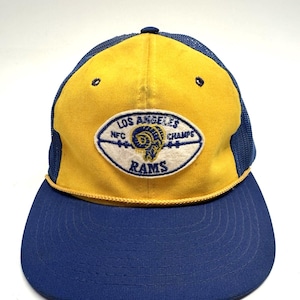 May include: A vintage Los Angeles Rams football team trucker hat. The hat features a yellow front panel with a blue mesh back and brim. The team logo is displayed on the front, with the text "LOS ANGELES NFC CHAMPS RAMS".