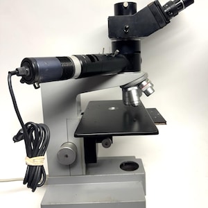 May include: A silver and black microscope with a black stage and a black and silver arm. The microscope has a black cord attached to it.
