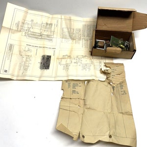 May include: A brown cardboard box with a torn piece of paper inside. The box contains a circuit board and other electronic components. The box is sitting on a white surface. There is a piece of paper with a circuit diagram on it.