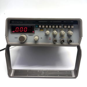 May include: A gray Instek function generator with a digital display showing "0.00". It has multiple knobs and buttons labeled "Coarse", "Frequency", "Fine", "Duty", "Offset", and "Amps". The device is on a gray stand.