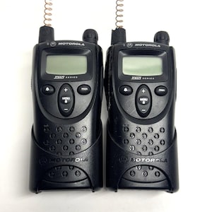 May include: Two black Motorola XTN series two-way radios. Each radio has a small screen, buttons, and a coiled antenna. The radios are in black holders and are set against a white background.