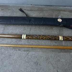 May include: A pool cue with a detailed carved design on the handle, accompanied by a separate cue tip and a black carrying case. The cue has white and gold accents. The case is black with a red interior.