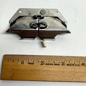 May include: A silver-colored metal object with a complex design, featuring two curved sections and a small, off-white cylindrical piece. A wooden ruler with inch markings and the text "Falcon" and "Auburn, Maine" is in the foreground.