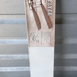 May include: A vintage model kit box for the Wright Flyer biplane. The box features a sepia-toned image of the aircraft and the text "WRIGHT FLYER". The box is white with brown and orange accents and is recommended for teens and adults with a 58-inch wing span.