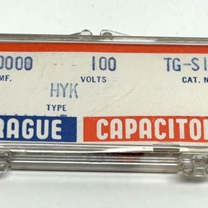 May include: A clear plastic case containing a Sprague capacitor. The case has a white label with black and blue text, including the words "SPRAGUE CAPACITORS." The label also displays technical specifications such as "100 VOLTS."