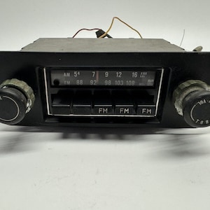 May include: A black vintage car radio with AM and FM bands. The radio has a dial with numbers from 54 to 108 and a series of buttons labeled "FM". The radio has two knobs on either side labeled "TONE".