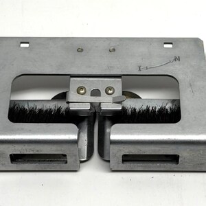 May include: A metal and brush component with a rectangular frame. The frame is silver and has a series of cutouts and mounting points. Black bristles are attached to the frame with wooden supports. The component has an arrow pointing to the letter "N".