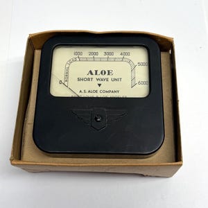May include: A black vintage "ALOE Short Wave Unit" meter with a white face and a needle indicator. The meter has a scale from 0 to 6000 and is housed in a cardboard box. The text "A.S. Aloe Company" is visible.